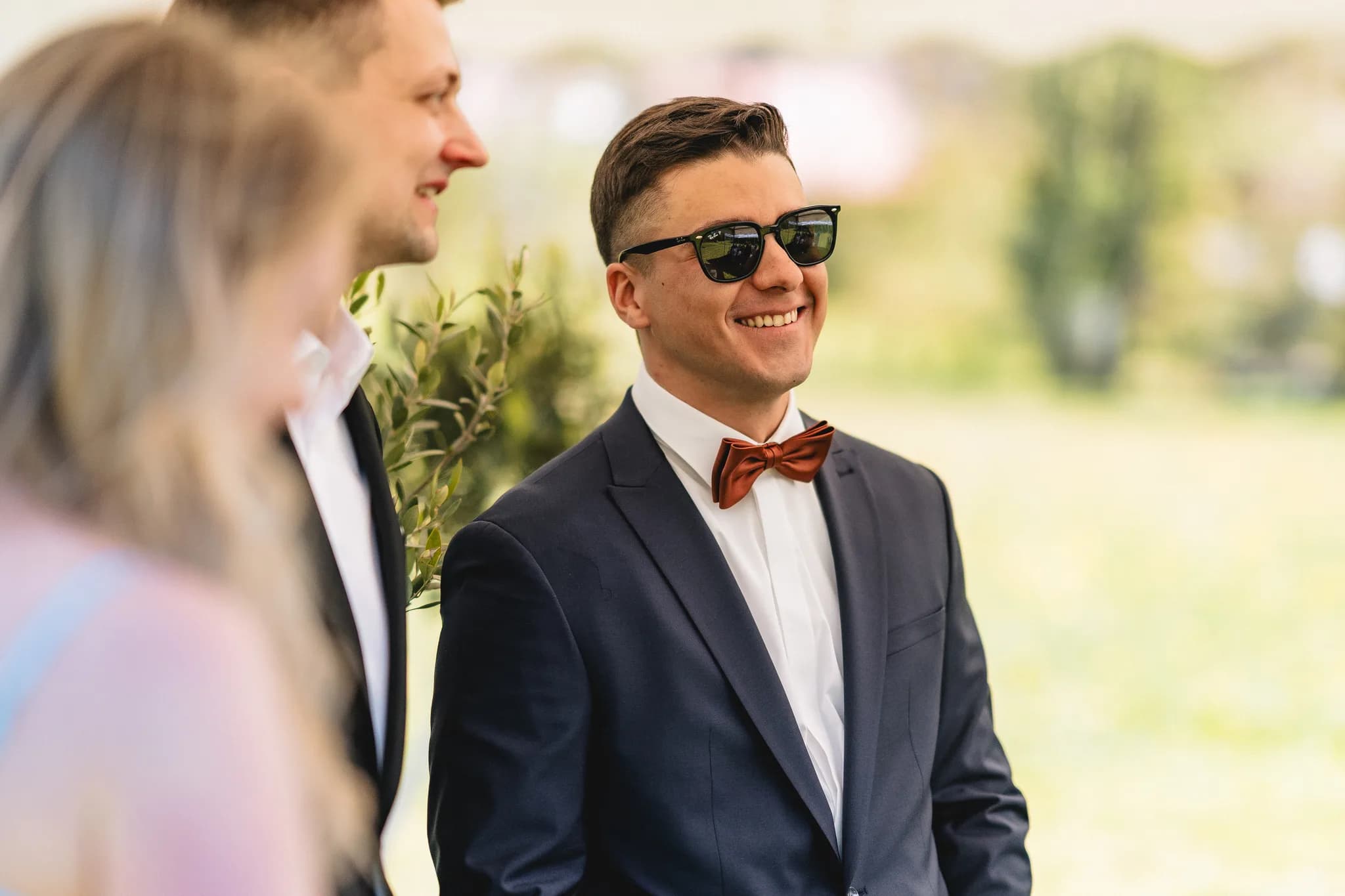 Der Bräutigam Ralf lächelt entspannt mit Sonnenbrille und Fliege während der freien Trauung auf der Wiese.