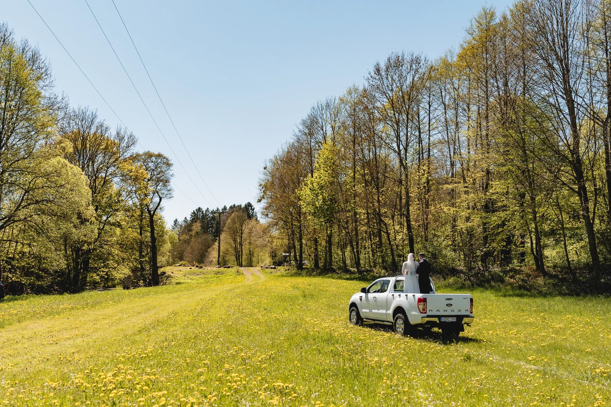 Brautpaar auf der Ladefläche eines Pickups auf einer sonnigen Blumenwiese in der Rhön an einem Maitag.