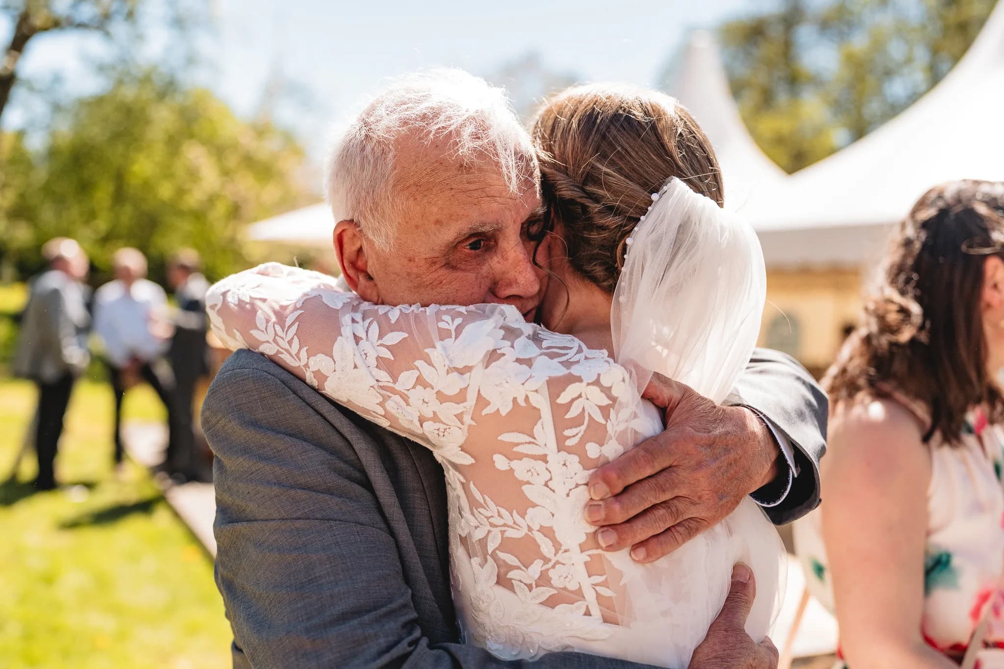 Emotionale Umarmung zwischen einer Braut in weißem Spitzenkleid und ihrem Vater bei einer Hochzeitsfeier im Grünen.