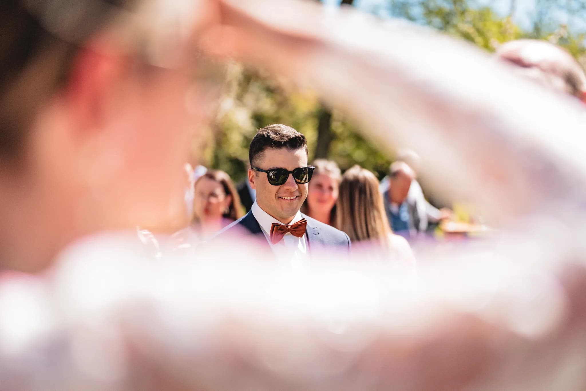 Bräutigam Ralf blickt mit Sonnenbrille und Fliege freudig durch einen Rahmen aus Blüten auf seine Braut bei der Trauung.
