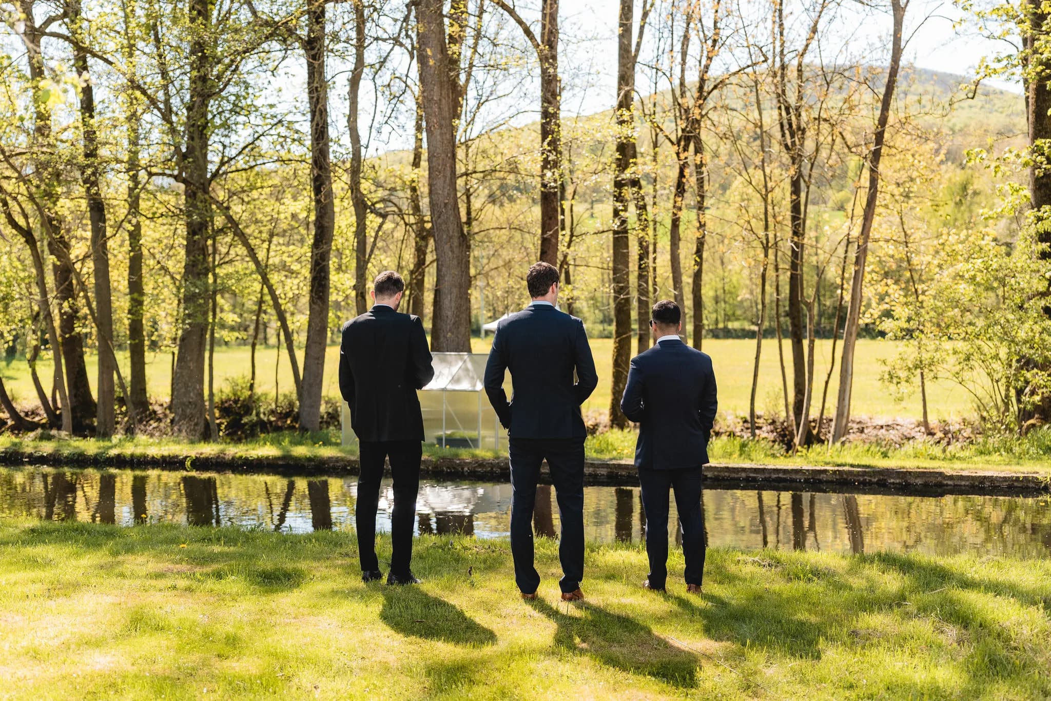 Drei festlich gekleidete Männer stehen entspannt an einem kleinen Bach auf einer grünen Wiese an der Ulstermühle.