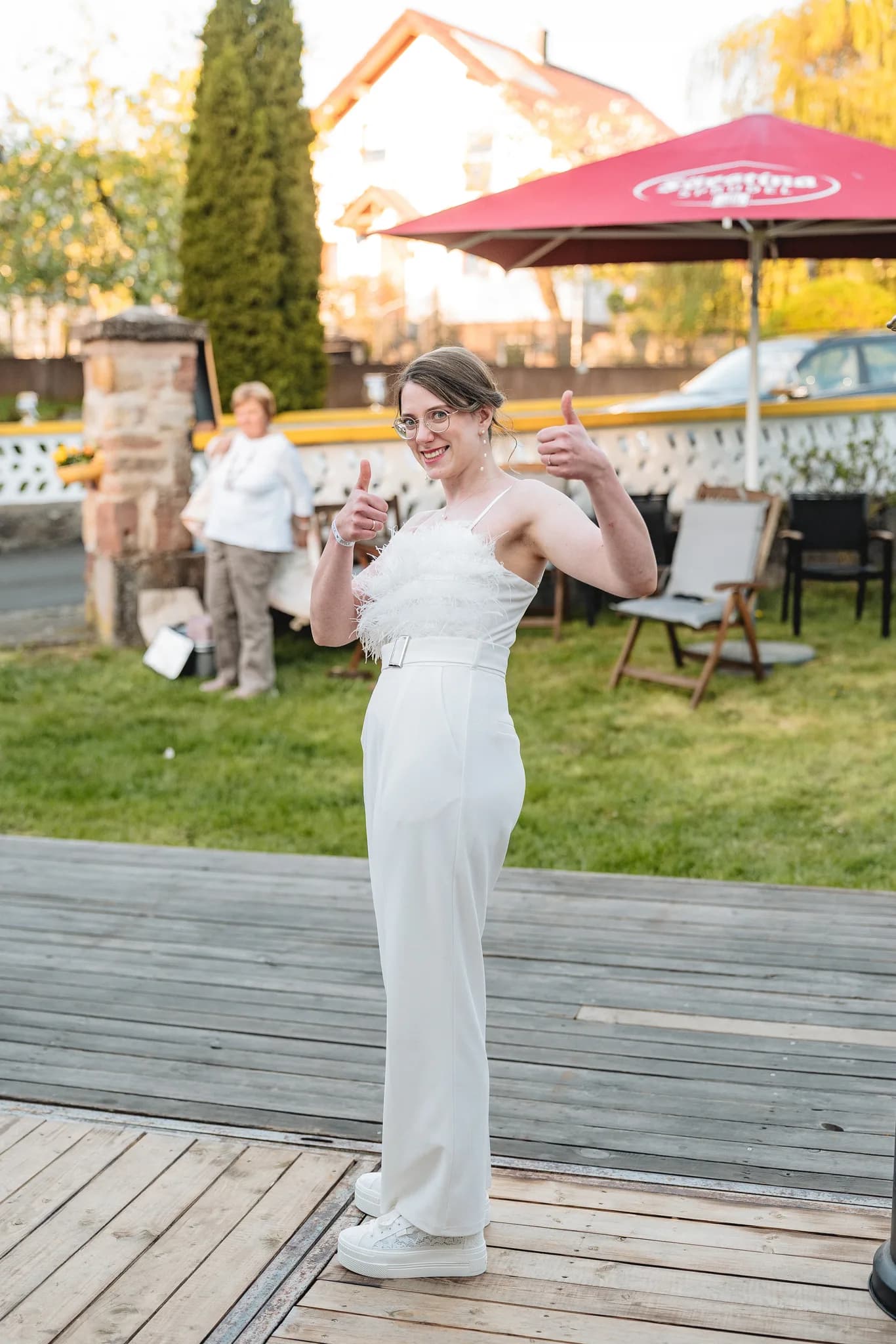 Die strahlende Braut Stefanie zeigt in ihrem modernen weißen Jumpsuit auf der Hochzeitsfeier im Garten beide Daumen hoch.