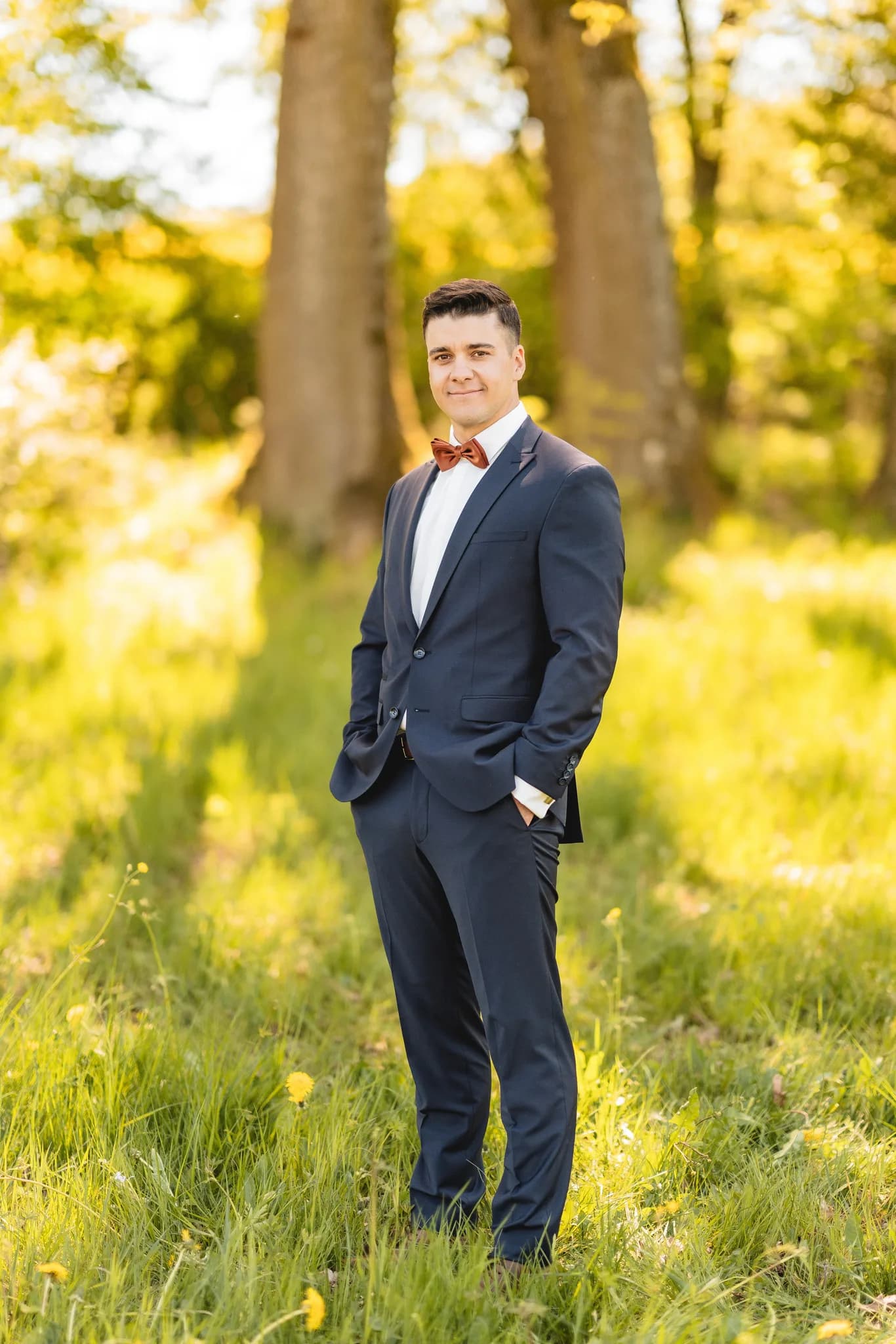 Bräutigam im dunkelblauen Anzug mit Fliege steht entspannt auf einer grünen Wiese an seinem Hochzeitstag.