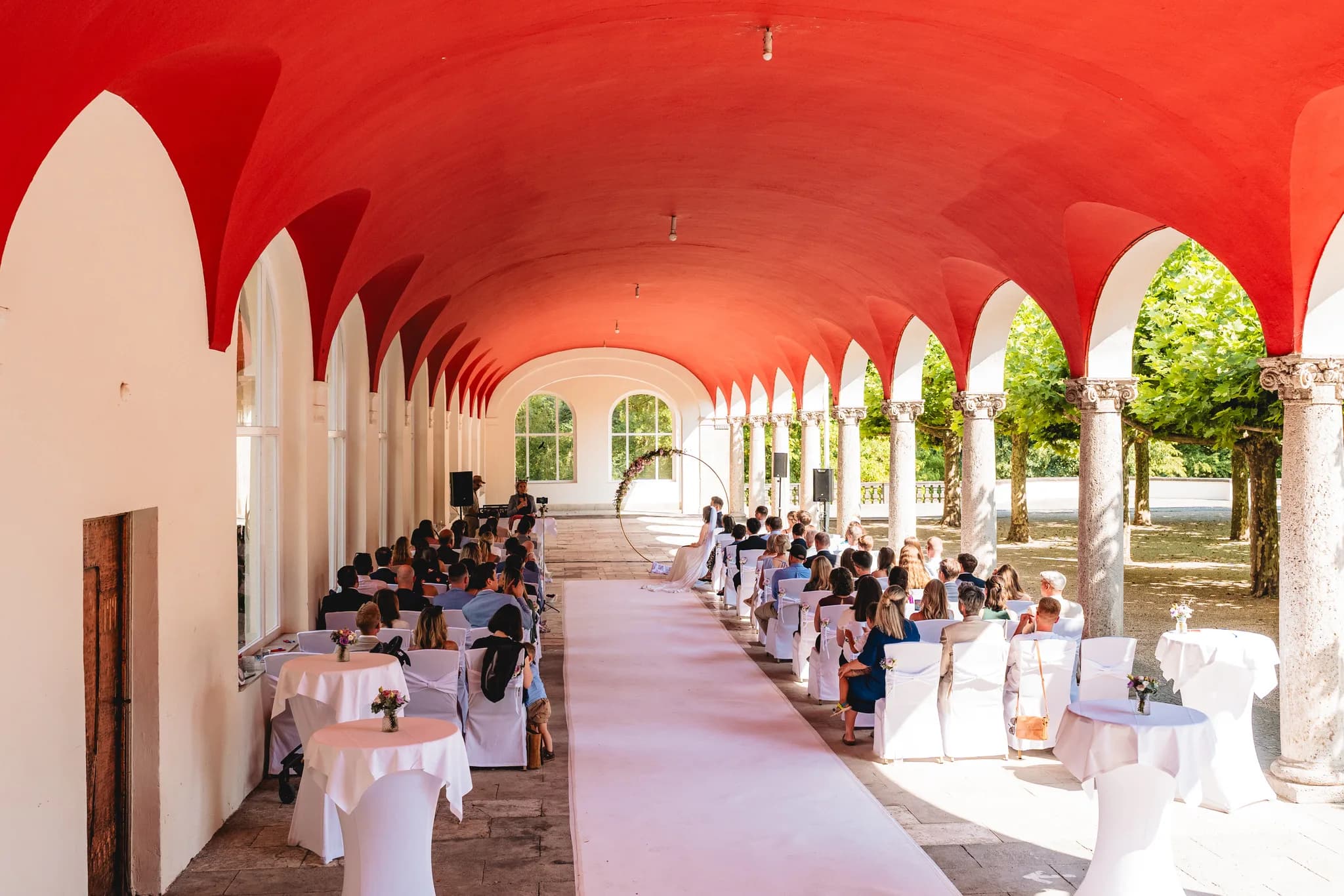 Gäste sitzen bei einer sonnigen Hochzeitszeremonie unter dem markanten roten Bogengang im Dolce by Wyndham Bad Nauheim.