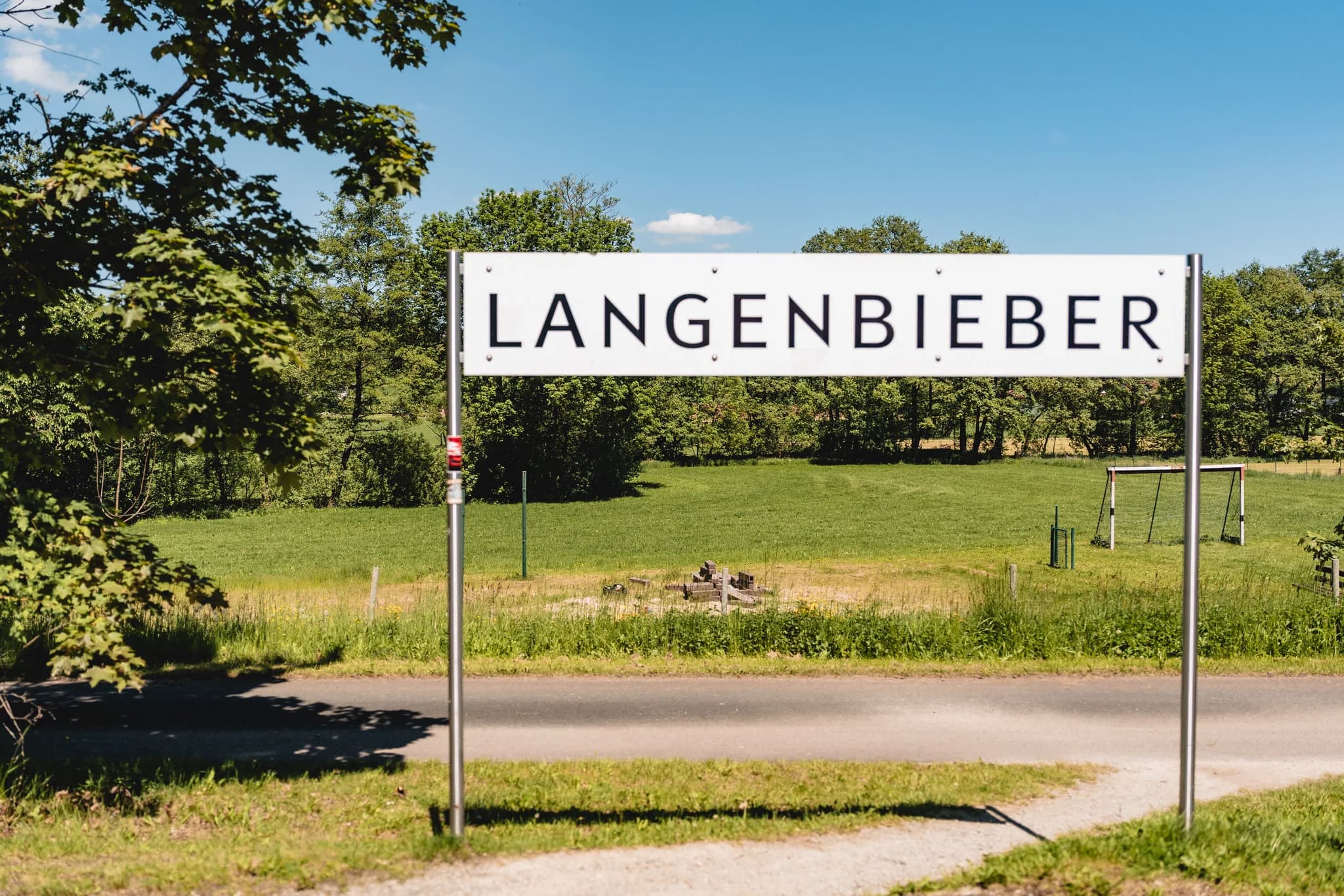 Ortsschild von Langenbieber an einer sonnigen Landstraße vor grünen Wiesen und Bäumen in Hofbieber.