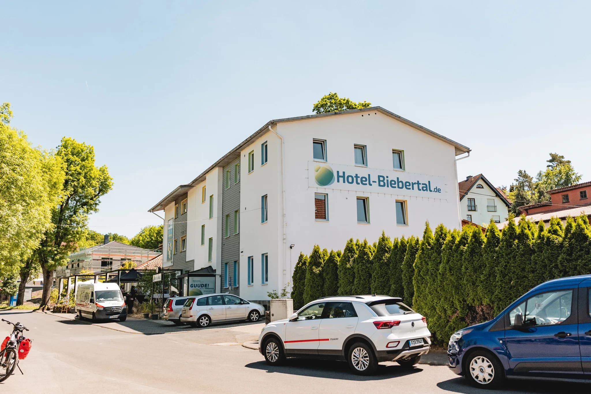 Außenansicht des Hotel-Biebertal in Hofbieber an einem sonnigen Tag mit geparkten Autos vor dem Gebäude.