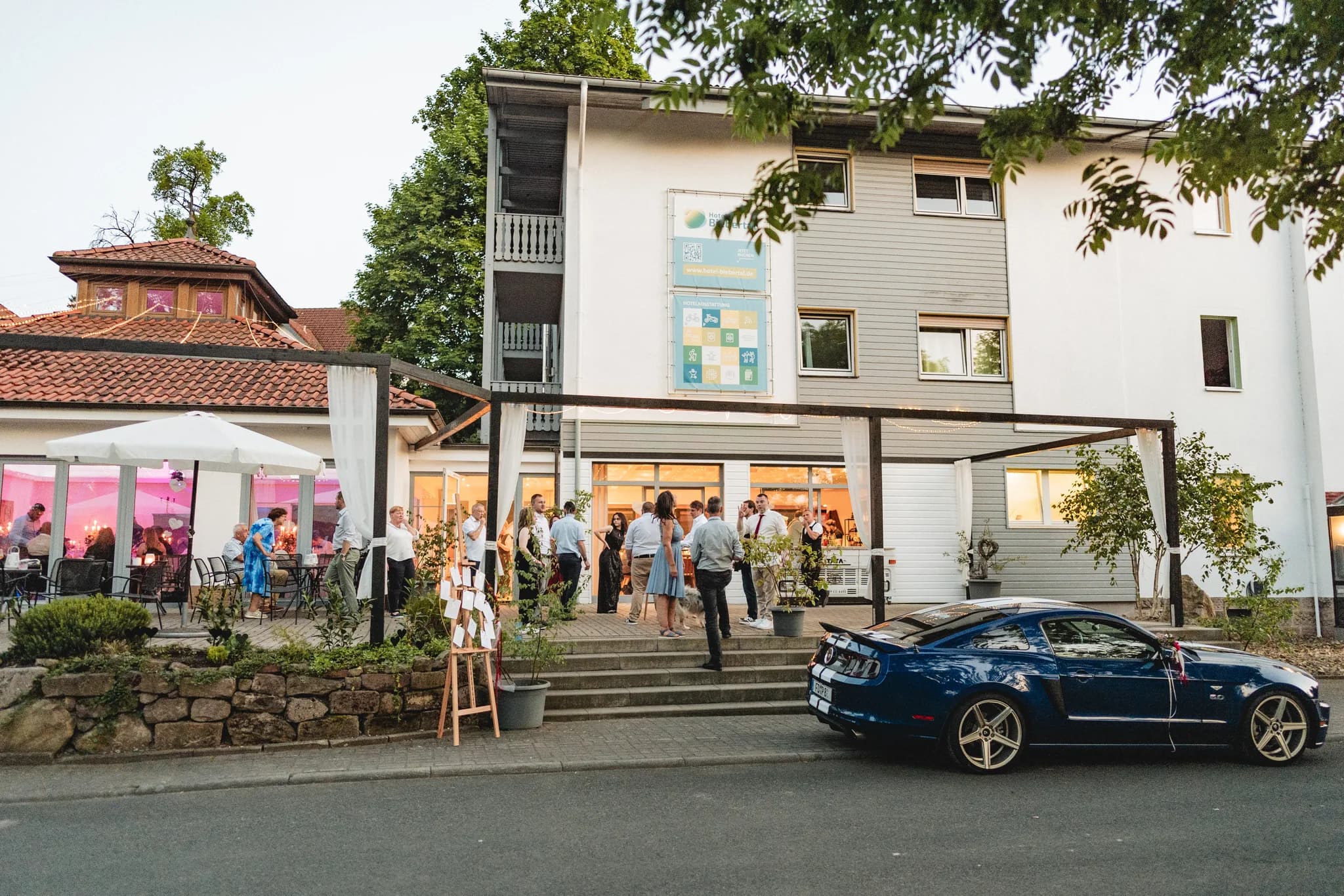 Hochzeitsgäste unterhalten sich abends auf der Terrasse vor einem Hotel in Hofbieber bei einem abgestellten Sportwagen.