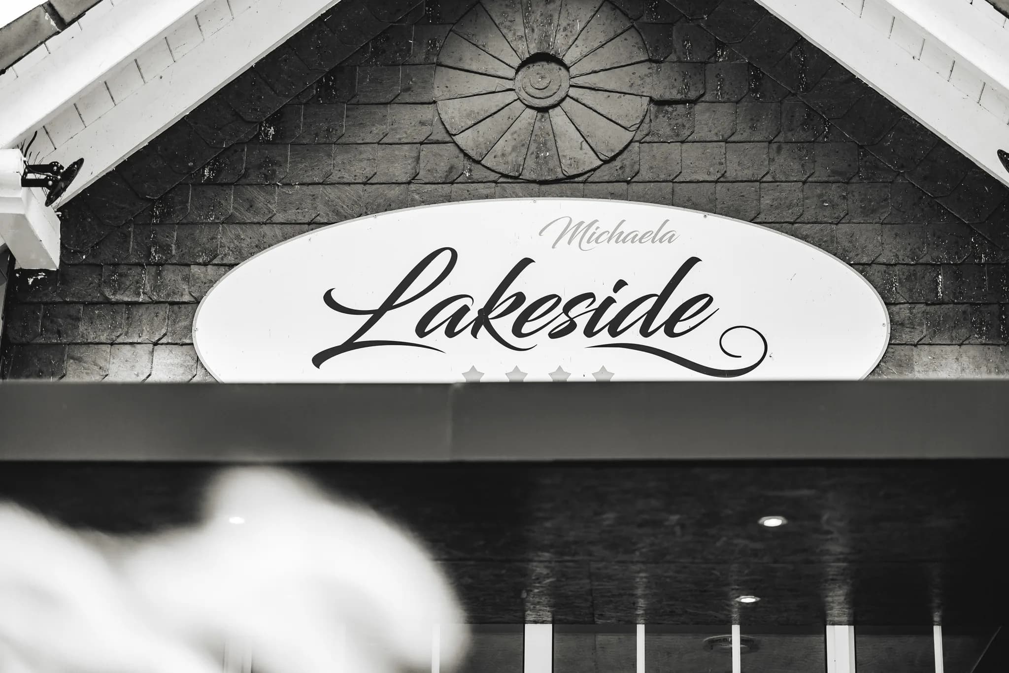 Schwarzes Firmenschild mit der Aufschrift Michaela Lakeside über dem Eingang eines Gebäudes in Schwarz-Weiß.