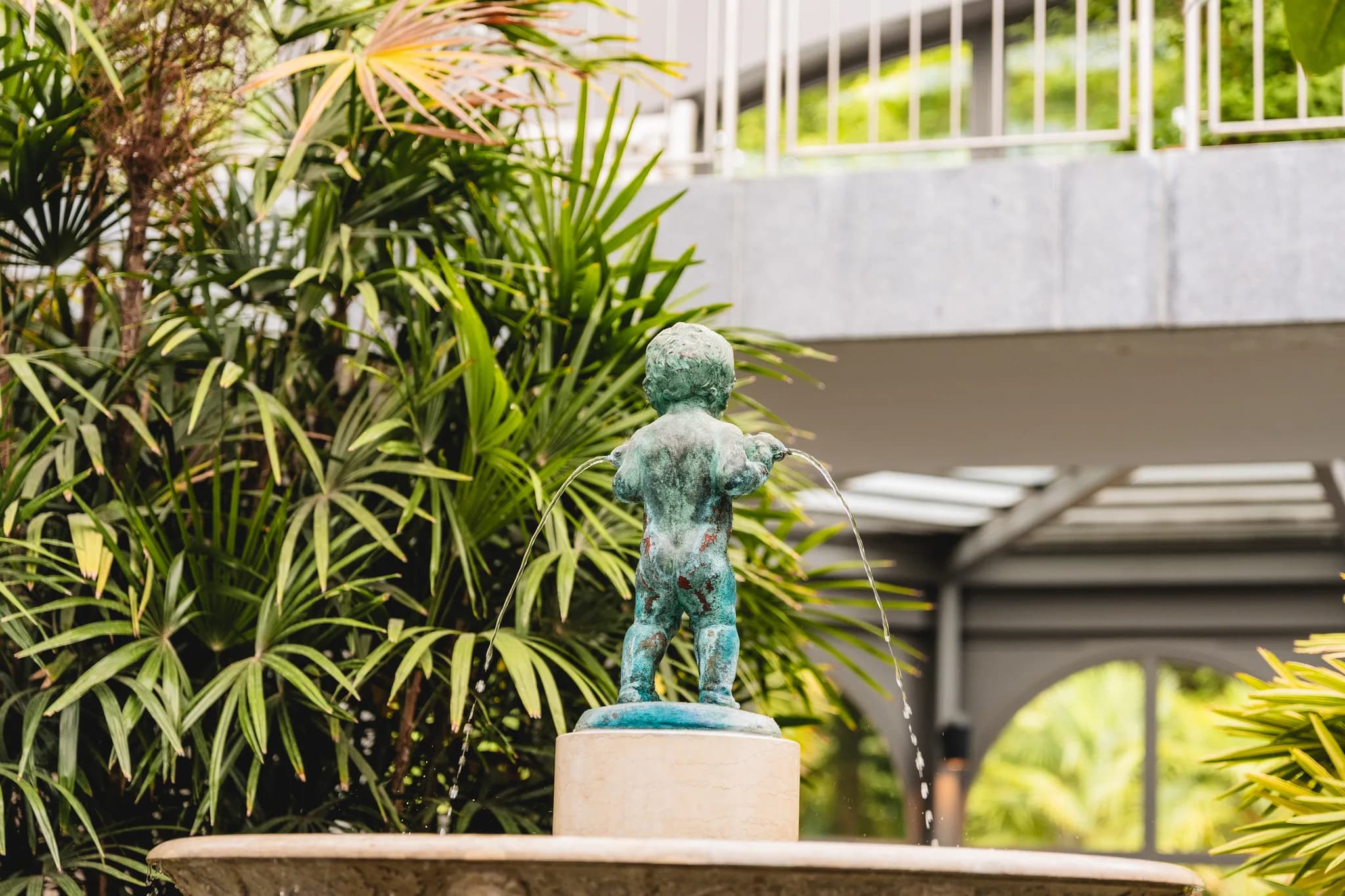 Kleine Putten-Brunnenfigur aus Bronze in einer üppig grünen Pflanzenkulisse im Palmengarten Frankfurt.