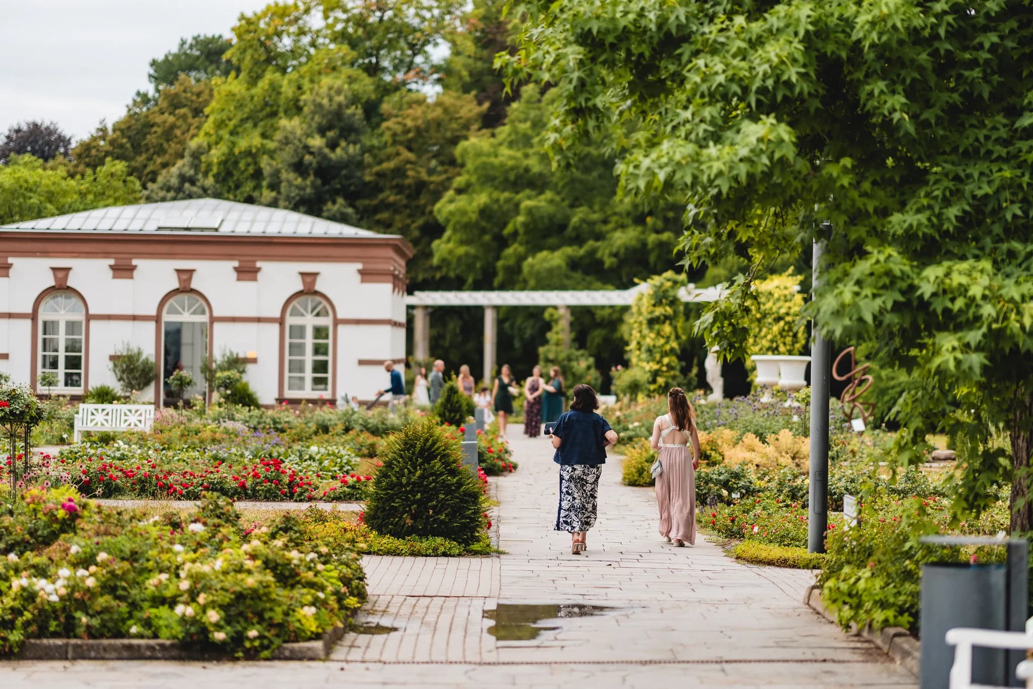 Hochzeitsgäste spazieren auf einem gepflasterten Weg durch die blühende Gartenanlage im Frankfurter Palmengarten.