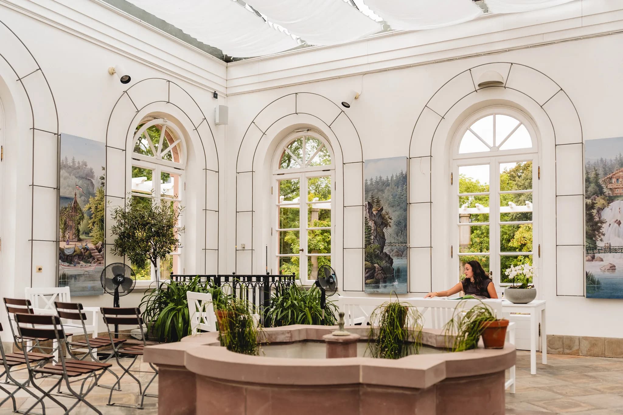 Eine Frau sitzt im hellen Gesellschaftshaus des Palmengartens Frankfurt an einem Tisch neben einem Steinbrunnen.