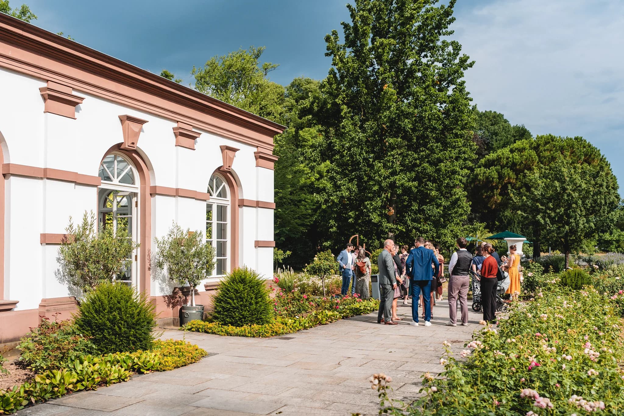 Hochzeitsgäste stehen vor einem historischen Gebäude im Palmengarten Frankfurt bei strahlendem Sonnenschein.