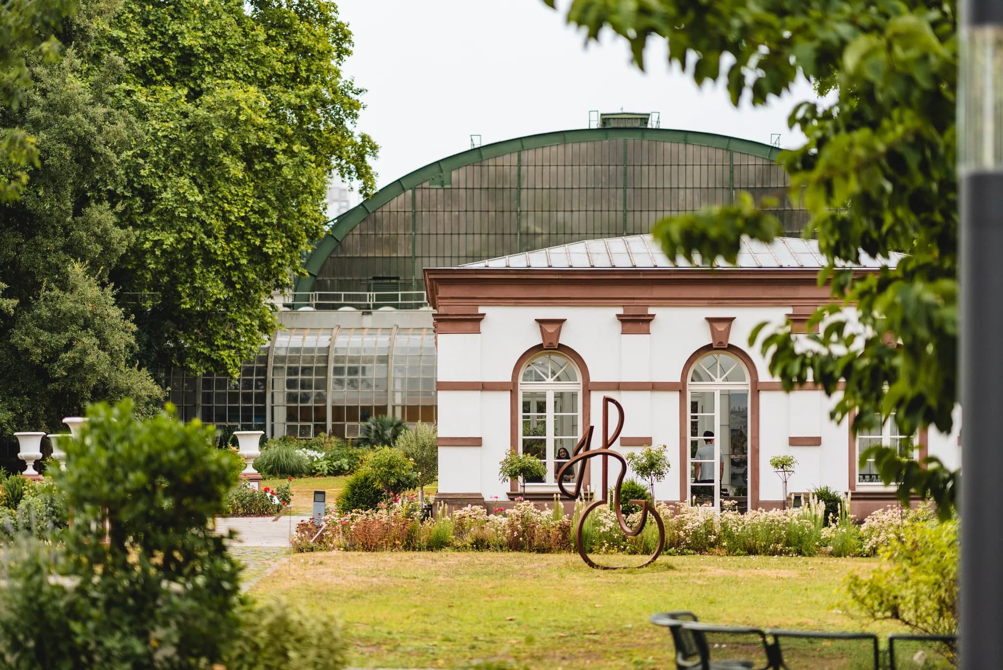 Blick auf das historische Gebäudeensemble im Frankfurter Palmengarten vor dem großen gläsernen Gewächshaus.
