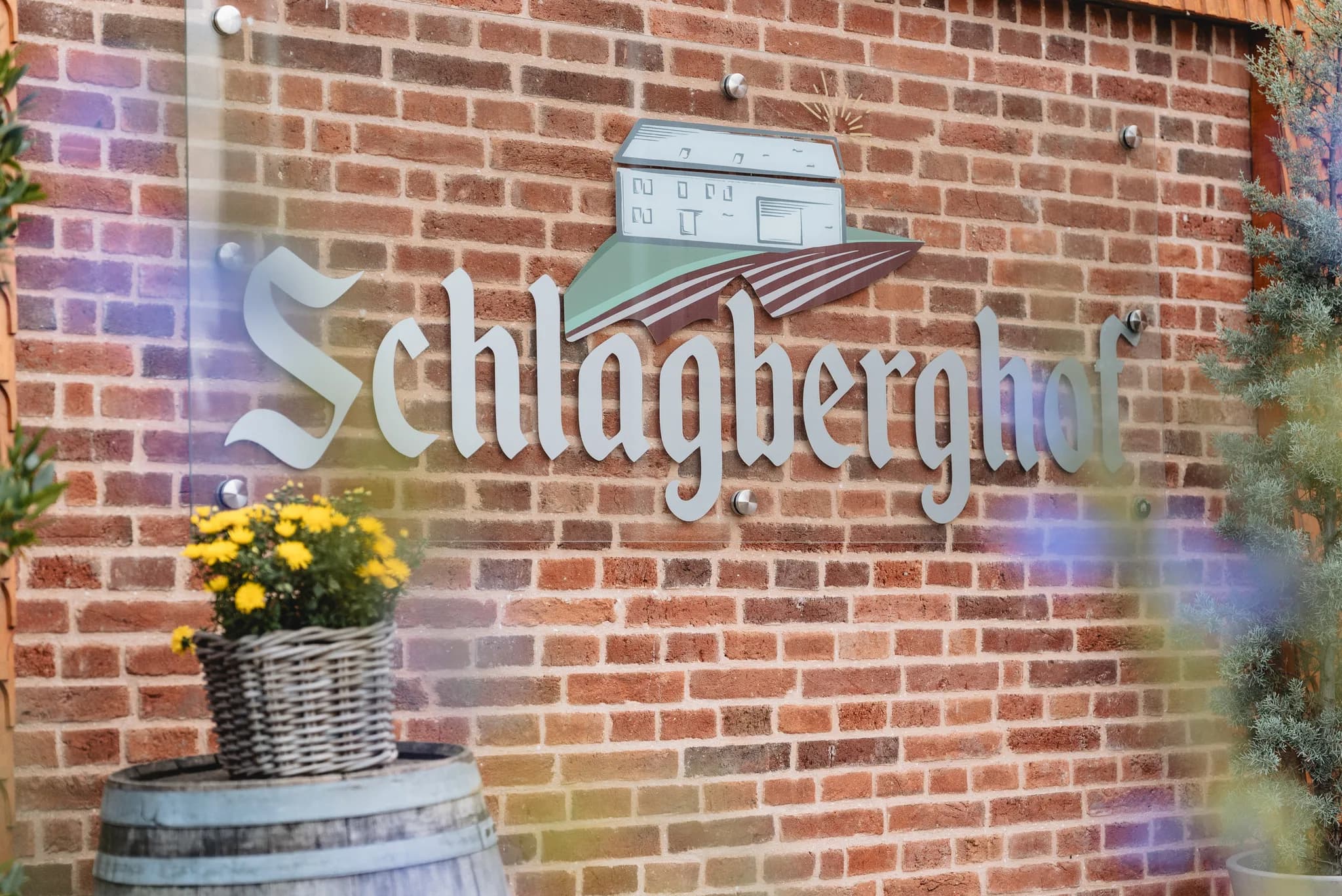 Schild mit der Aufschrift Schlagberghof vor einer roten Backsteinwand in Kleinlüder mit einem Blumenkorb auf einem Fass.