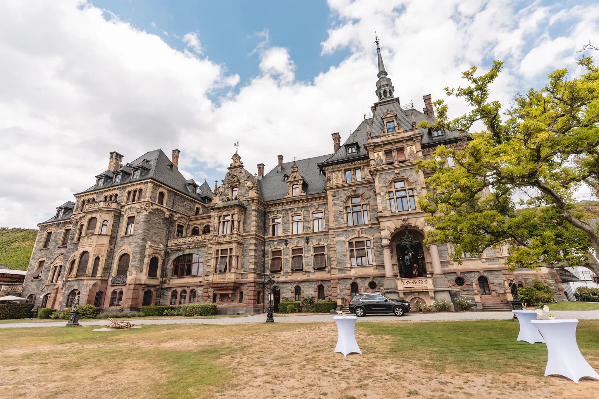 Historisches Schloss Lieser an der Mosel mit Steinfassade und weißen Stehtischen auf dem gepflegten Gartenareal.
