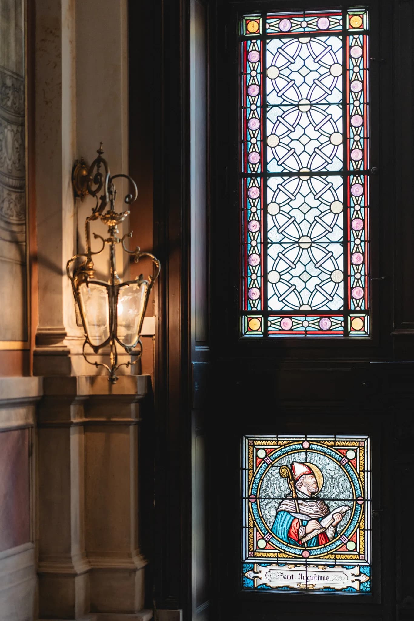 Antike Wandlampe neben einem farbigen Bleiglasfenster mit Heiligenfigur in einer Kirche in Lieser an der Mosel.