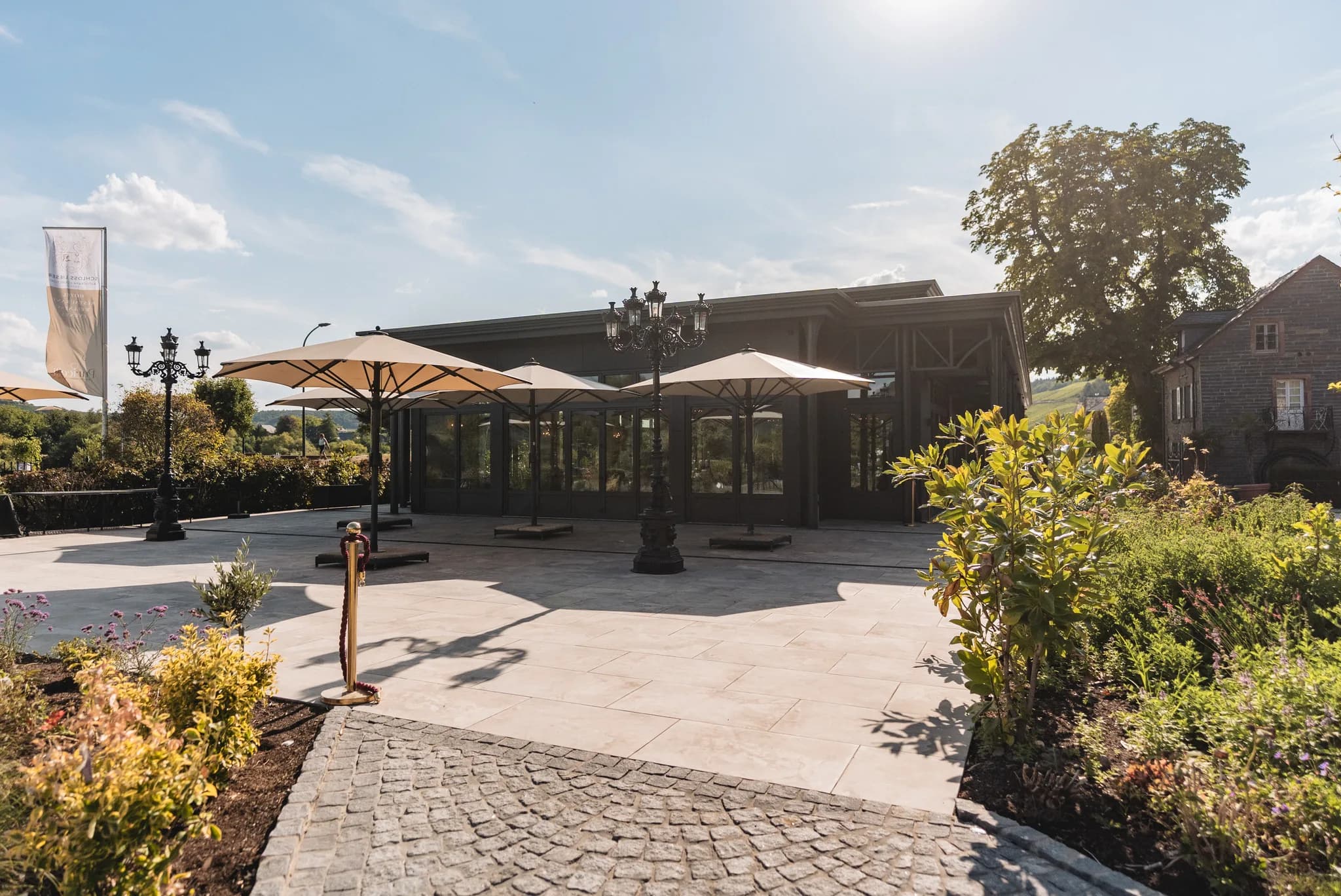 Sonnige Terrasse vor dem modernen Glasgebäude von Schloss Lieser an der Mosel mit großen Sonnenschirmen.