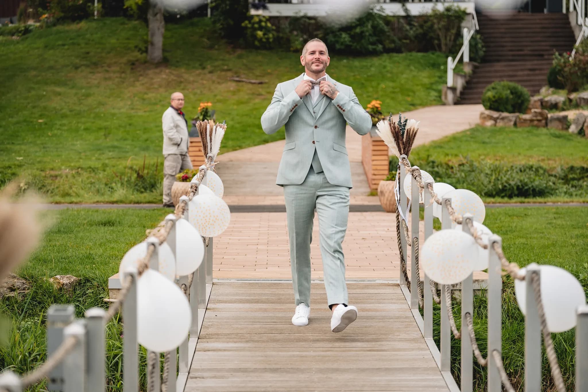 Bräutigam Daniel richtet sich auf einer mit Ballons geschmückten Holzbrücke im Lakeside Resort Michaela die Fliege.
