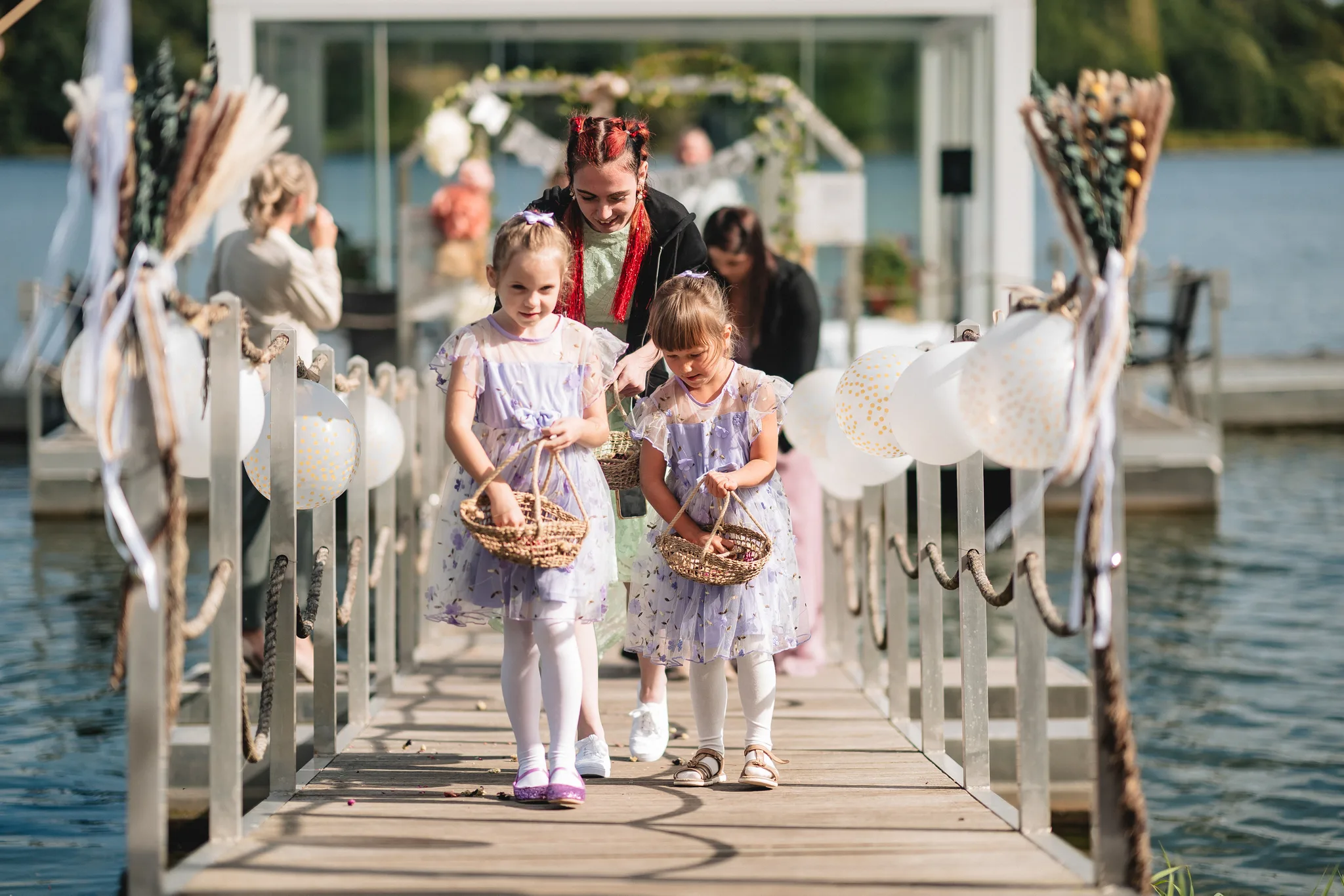 Zwei Blumenmädchen in lila Kleidern schreiten mit Körbchen über den Steg bei der Hochzeit im Lakeside Resort Michaela.