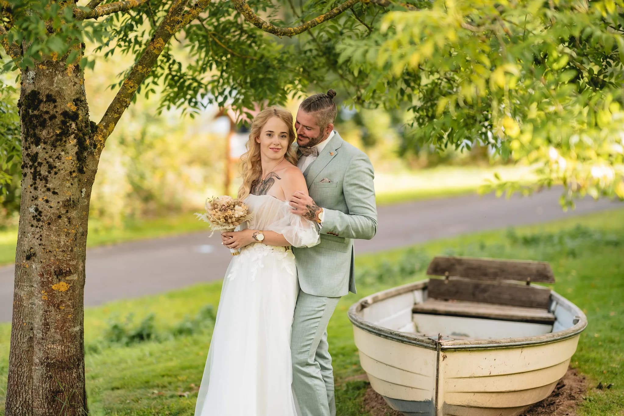 Brautpaar Peggy und Daniel stehen bei ihrer Hochzeit im Lakeside Resort Michaela an einem alten Holzboot unter Bäumen.
